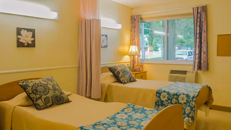 A bright and cozy room with two single beds, each with beige bedspreads and blue floral-patterned throws and pillows. A window with floral curtains lets in natural light, and a bedside table with a lamp and a small decorative item is placed between the beds. The walls are light-colored with a small flower painting and a curtain divider between the beds.