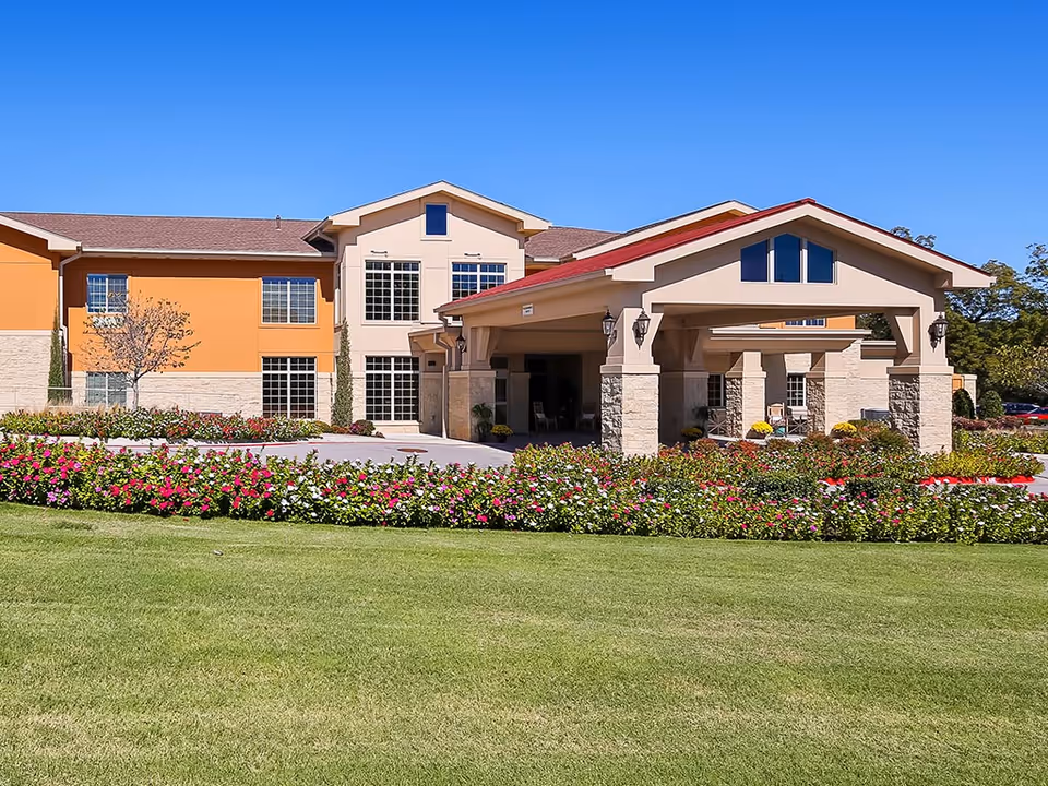 Front entrance of a two-story senior living building with a covered porte-cochere, manicured lawn, and colorful flower beds.