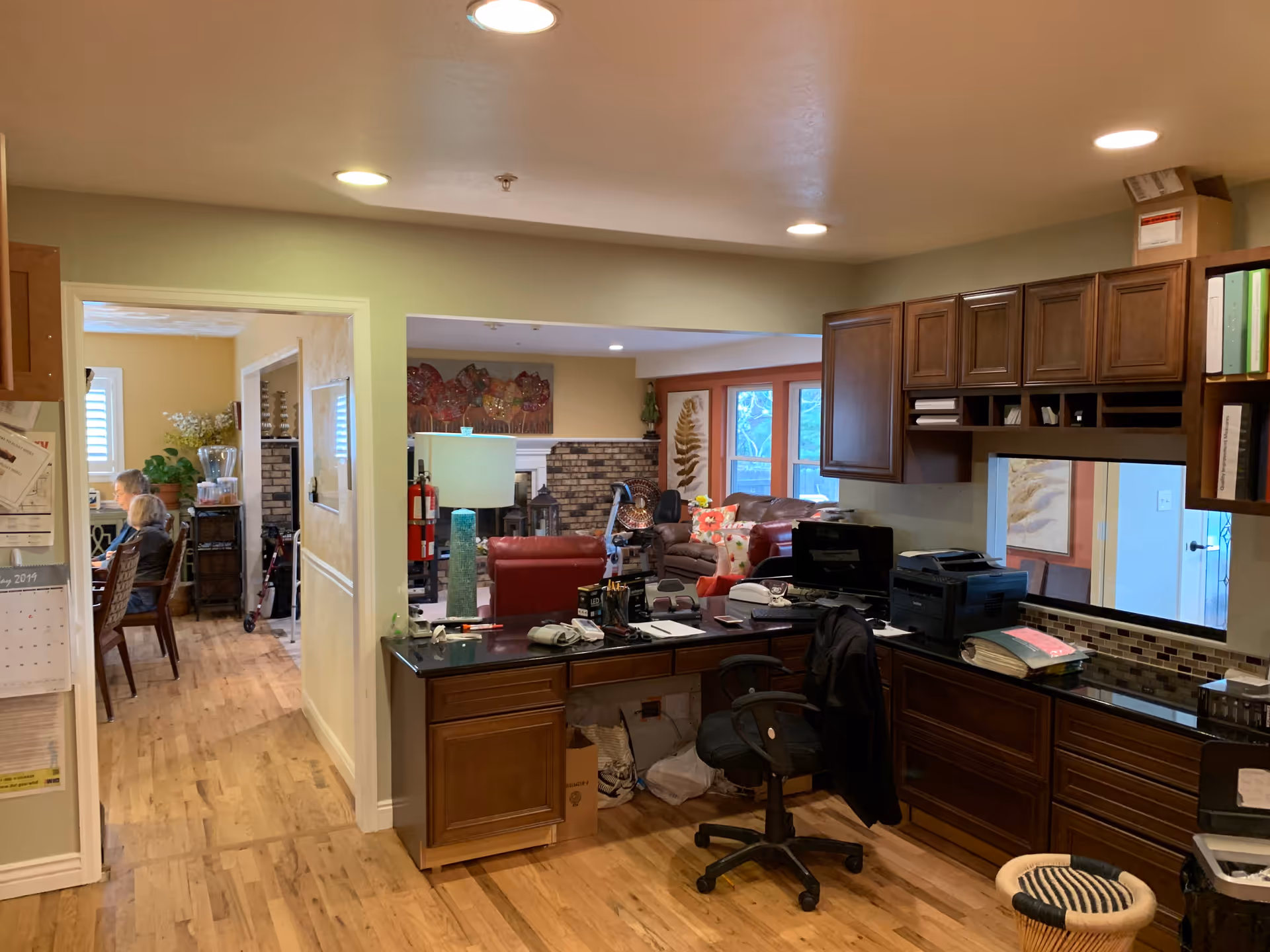 Interior view of Serenity House Assisted Living Centennial showing a reception or office area with wooden cabinets, a desk with office supplies, and a chair. In the background, there is a living room with a brick fireplace, leather sofas, and decorative wall art. To the left, a hallway leads to a dining area where two elderly people are seated at a table.