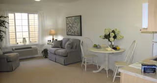 A cozy senior living room with a gray couch, a matching armchair, a small round dining table with two white chairs, a vase with flowers on the table, a lamp on a side table, and a large window with white curtains letting in natural light.