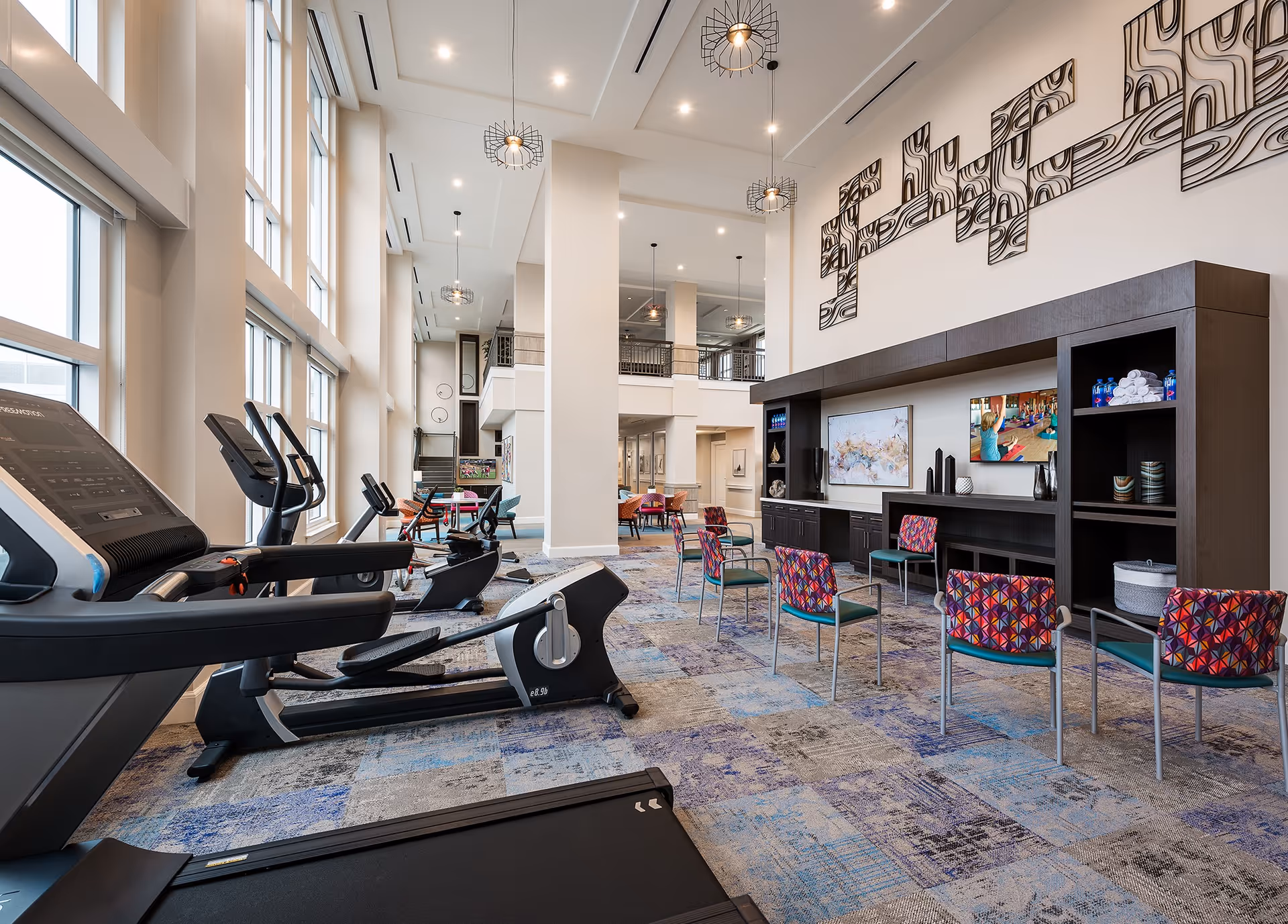 A spacious fitness and activity room with large windows letting in natural light. The room features several exercise machines including treadmills and elliptical trainers on the left side. On the right side, there are colorful chairs arranged in front of a large dark wood entertainment center with two flat-screen TVs and decorative items. The room has high ceilings with modern pendant lights and a patterned carpet in shades of blue and gray. In the background, there is a seating area with tables and chairs and a staircase leading to an upper level.