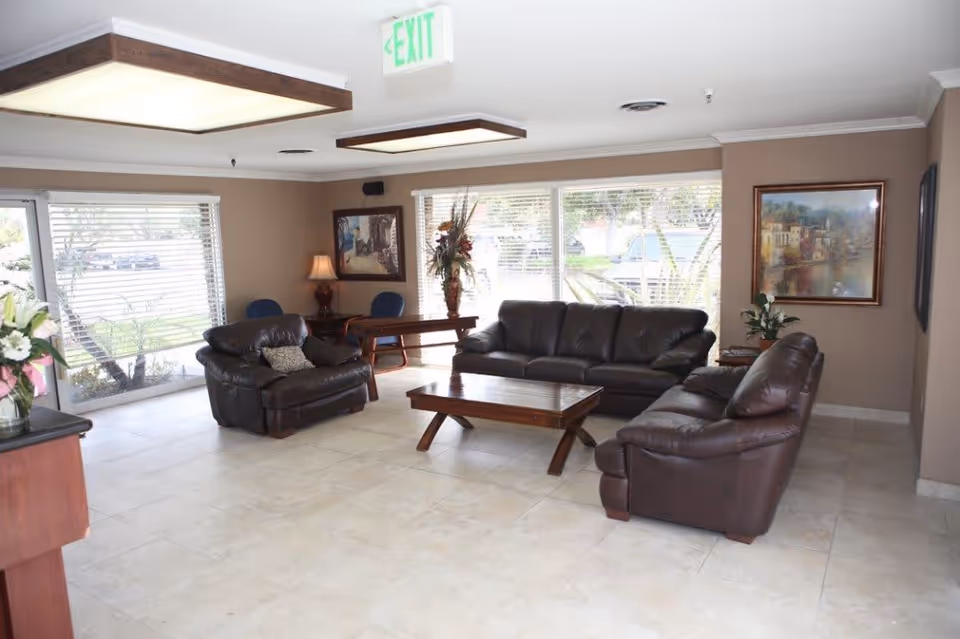 A well-lit living room area with three dark brown leather sofas arranged around a wooden coffee table. Large windows with blinds allow natural light to fill the room. The walls are beige and decorated with framed paintings and a floral arrangement on a side table. There is an exit sign on the ceiling near the center of the room.