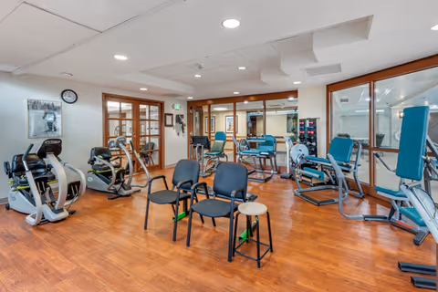 A fitness room with various exercise equipment including stationary bikes, weight machines, and chairs arranged on a wooden floor. The room has large mirrors on one wall and glass doors leading to another area.