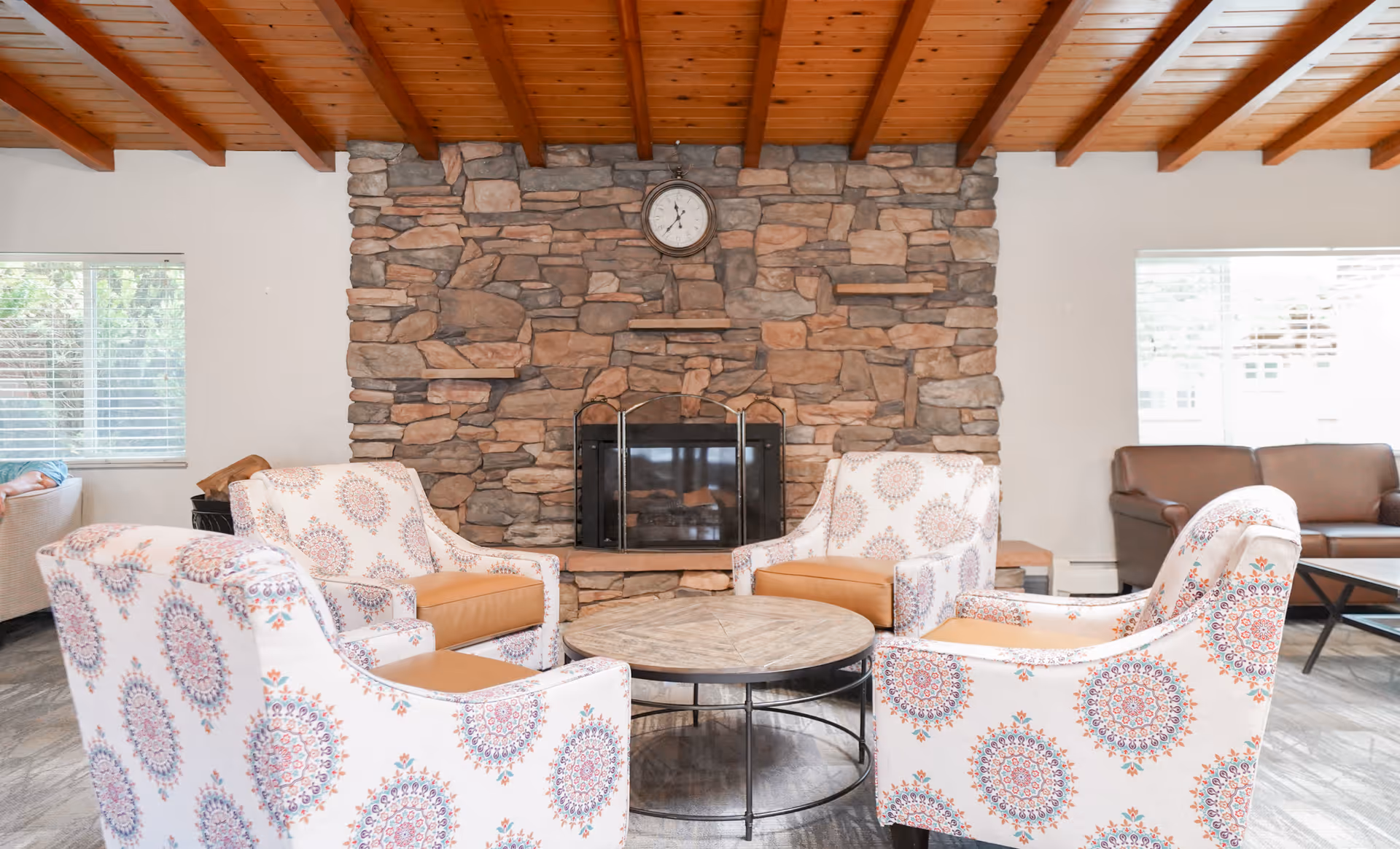 A cozy living room area with four patterned armchairs arranged around a round wooden coffee table. Behind the chairs is a stone fireplace with a clock mounted above it. The room has a wooden ceiling with exposed beams and large windows letting in natural light.