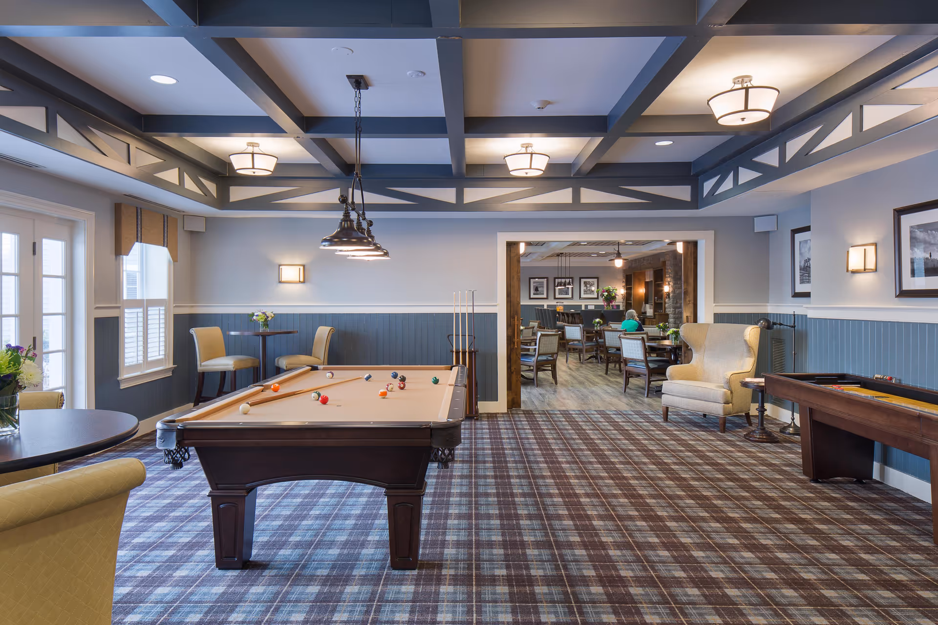 A spacious recreational room in a senior living facility featuring a pool table with balls arranged on it, a shuffleboard table to the right, and several seating areas with chairs and tables. The room has a patterned carpet, light blue wainscoting on the walls, and a coffered ceiling with multiple light fixtures. In the background, there is an adjoining room with more tables and chairs, and a person seated at one of the tables.