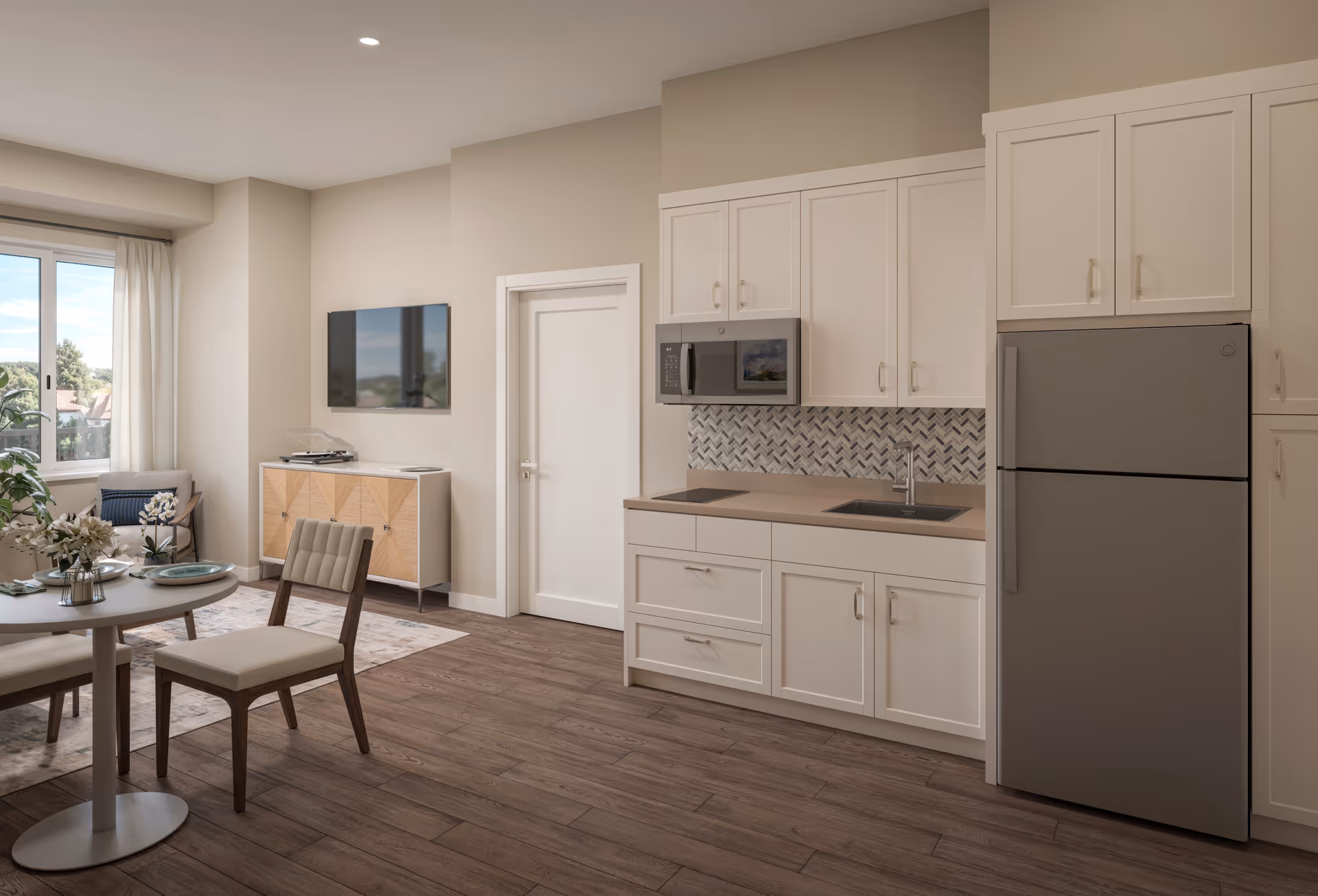 Open-plan apartment interior showing a modern kitchenette with white cabinets, stainless refrigerator and microwave, and an adjacent dining table and seating near a window.