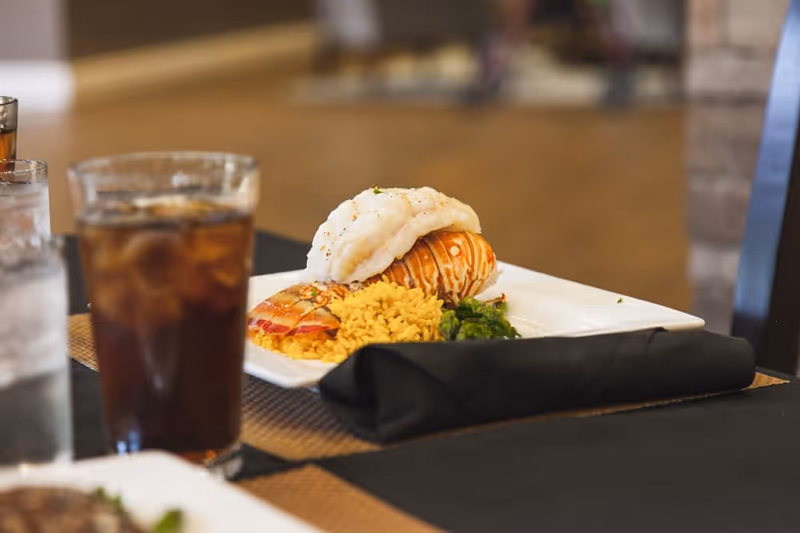 A close-up of a plated meal featuring a lobster tail, yellow rice, and green vegetables on a white square plate, with a black cloth napkin beside it and a glass of iced beverage in the background on a dining table.