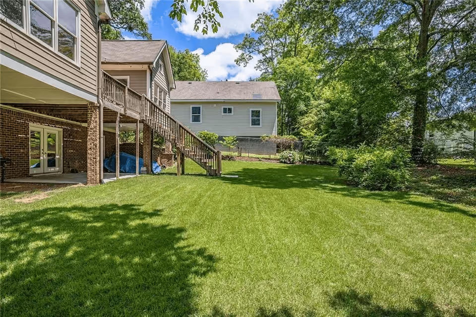 Green backyard with a raised wooden deck and staircase attached to a house, lawn and trees with a neighboring house in the background.