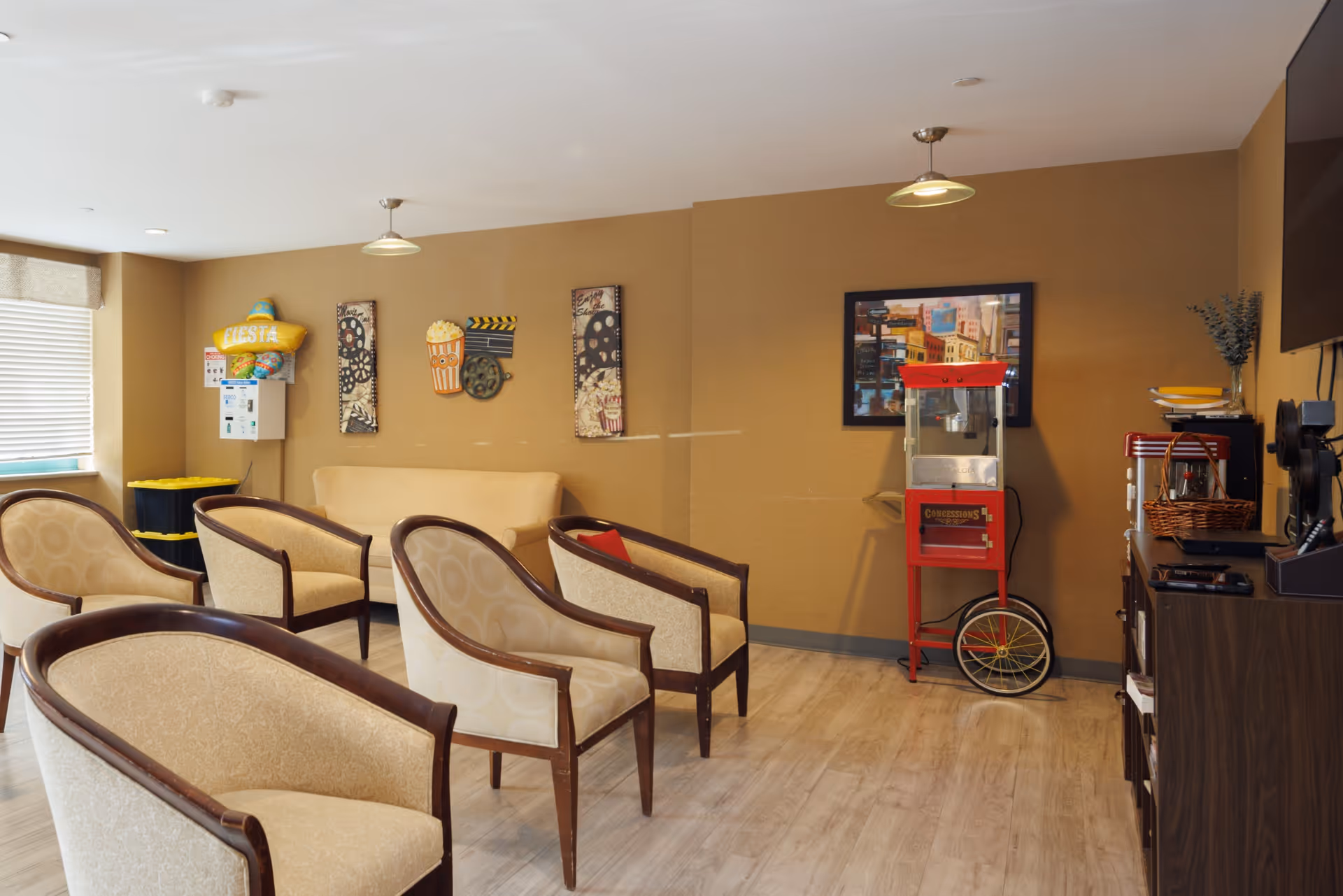 A cozy lounge area with several beige upholstered chairs with wooden frames arranged facing a popcorn machine on a red cart. The walls are decorated with movie-themed artwork and a framed picture. There is a beige couch against the wall and a window with blinds letting in natural light. A TV is mounted on the right wall above a dark wooden cabinet with various items on top.