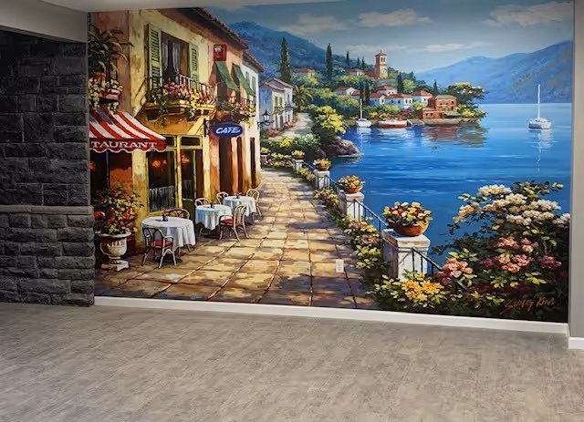 A large mural painting on an interior wall depicting a scenic waterfront village with a stone walkway, outdoor café tables with white tablecloths, colorful flowers in pots, and boats on calm blue water under a partly cloudy sky.
