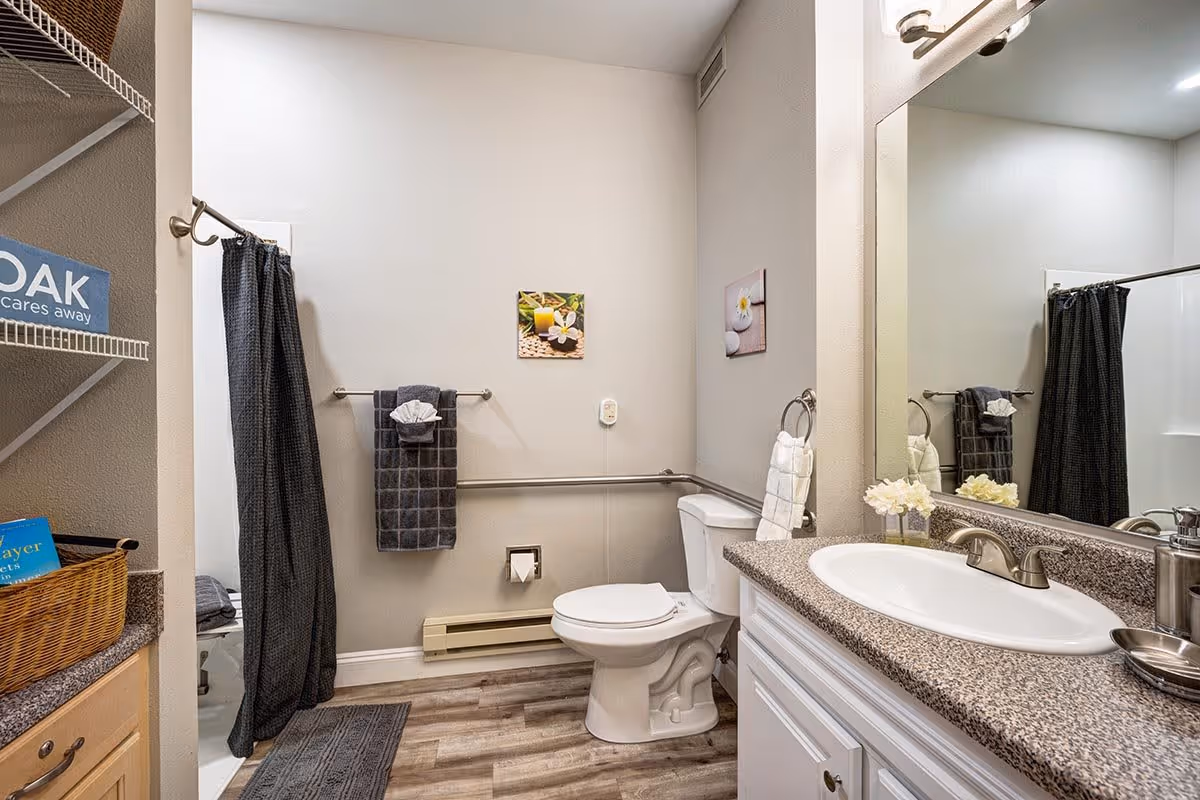 A clean and modern bathroom with a white toilet, a sink with a granite countertop, a large mirror, and a shower with a dark curtain. The walls are light-colored with two small decorative pictures hanging. There are towels hanging on racks and a small basket on a shelf to the left.