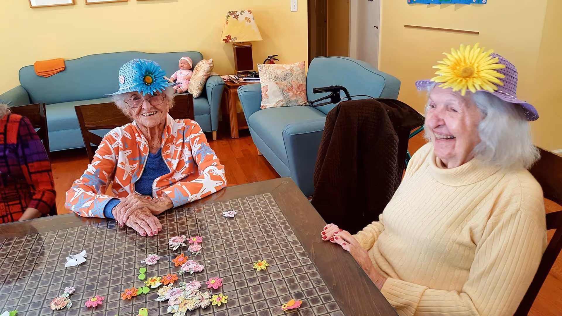 Two elderly women wearing decorative hats smile while sitting at a table with scattered craft flowers in a cozy communal room.