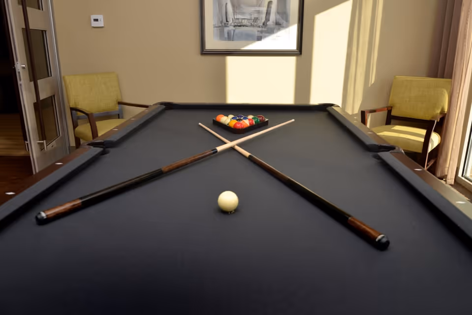 A pool table with a black felt surface, two crossed pool cues, a white cue ball, and a rack of colorful billiard balls at the far end. The room has beige walls, two yellow cushioned chairs on either side of the table, a framed picture on the wall, and sunlight streaming in through a window with curtains.