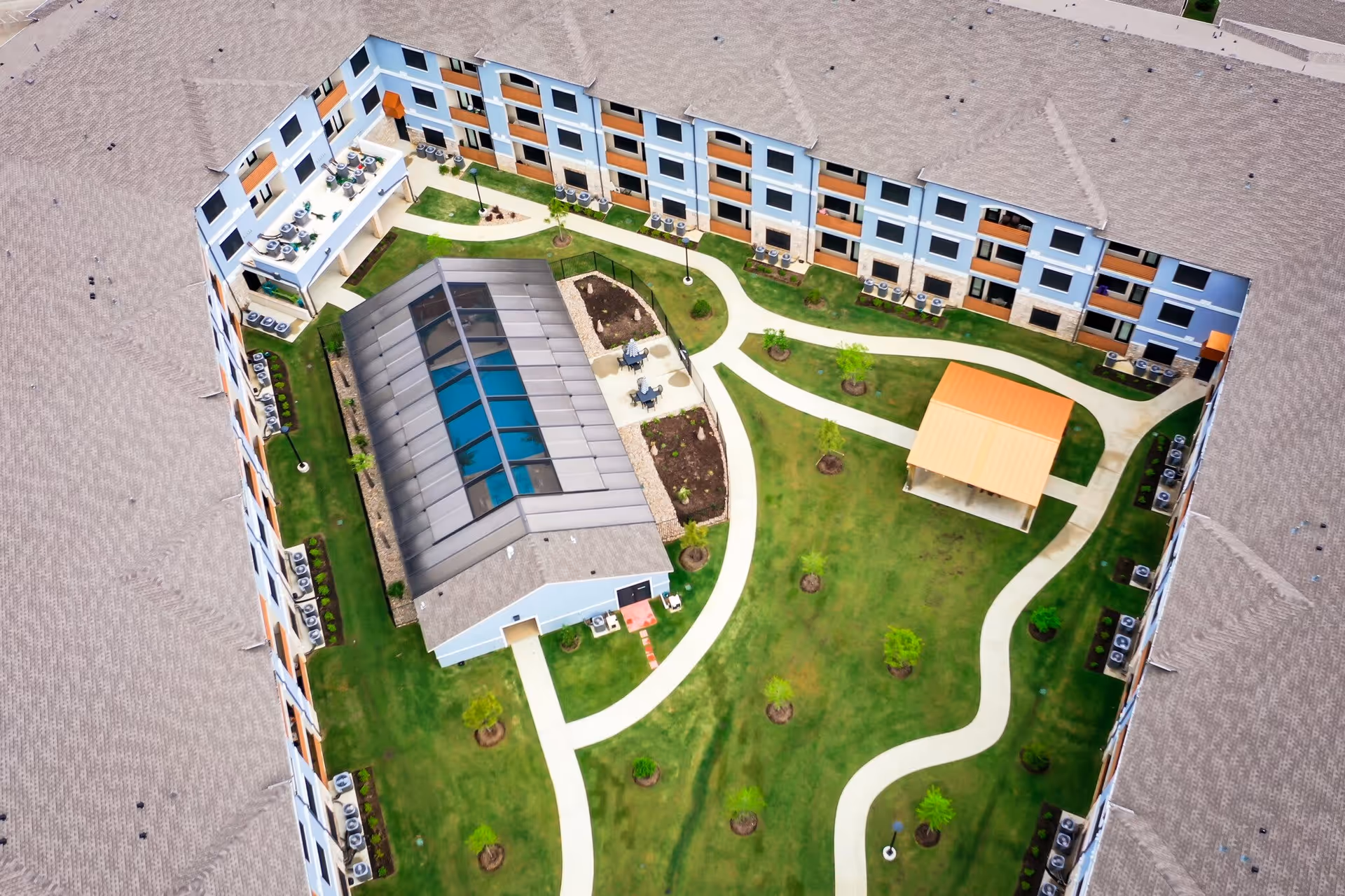 Aerial view of a senior living facility courtyard with a central building featuring a glass roof, surrounded by a three-story building. The courtyard has green lawns, walking paths, small trees, a gazebo with an orange roof, and outdoor seating areas.