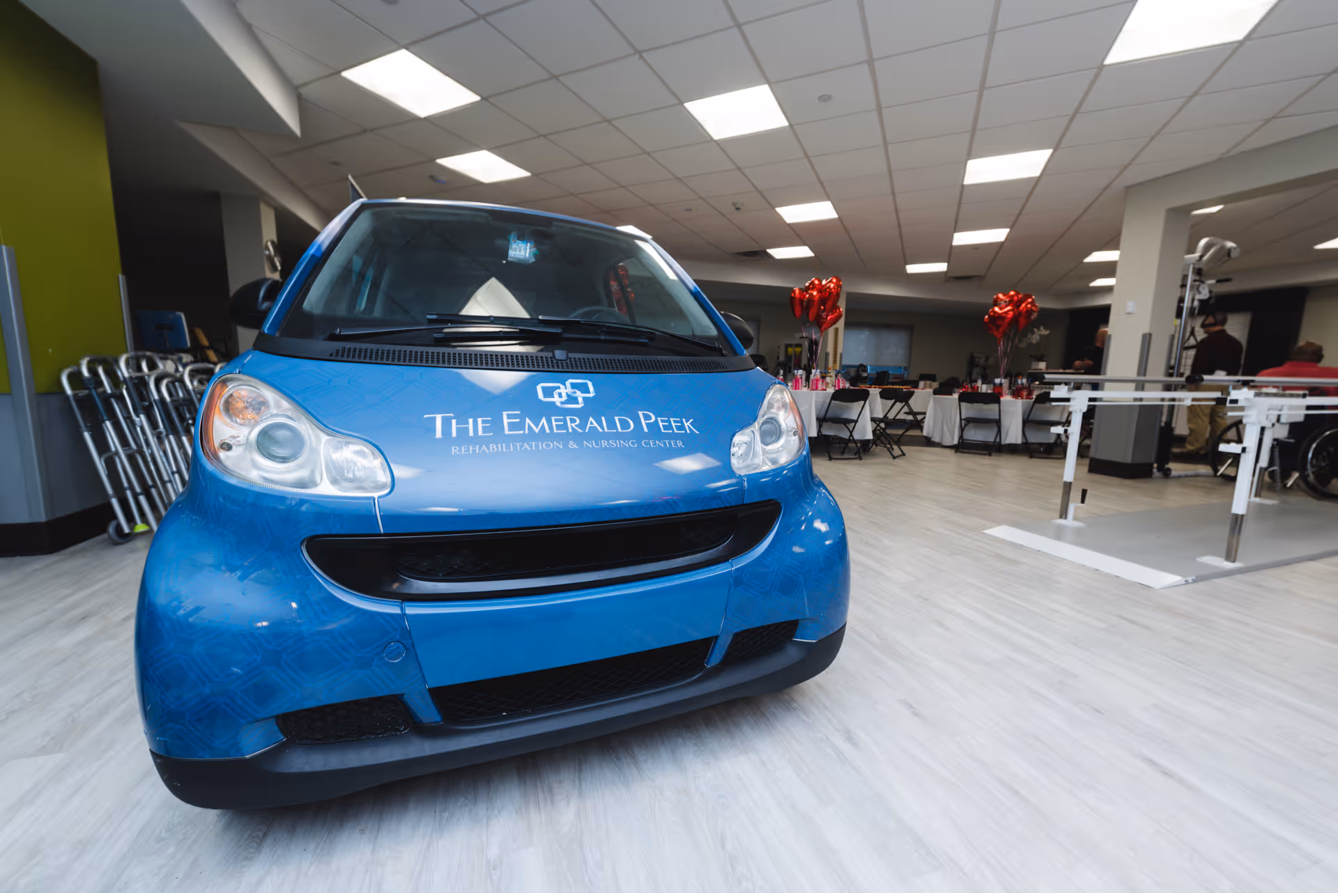 A small blue car with the logo and name 'The Emerald Peek Rehabilitation & Nursing Center' on the hood is parked inside a spacious, well-lit room with light-colored flooring. In the background, there are tables with red heart-shaped balloons, chairs, and some medical or rehabilitation equipment. Several walkers are lined up against a wall on the left side.