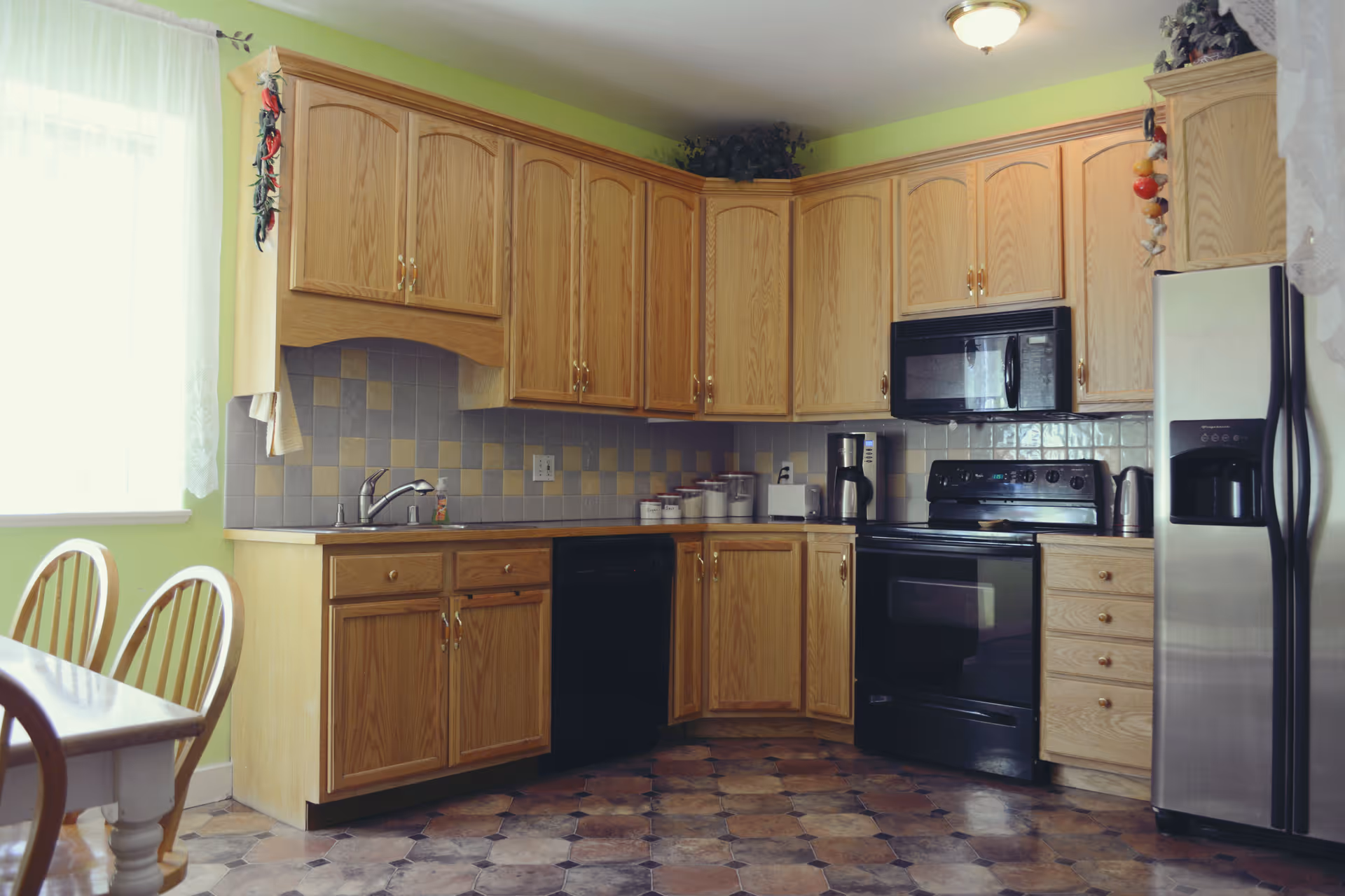 A kitchen with wooden cabinets, a black stove and microwave, a stainless steel refrigerator, a coffee maker, a toaster, and a sink under a window with sheer white curtains. There is a dining table with wooden chairs on the left side.