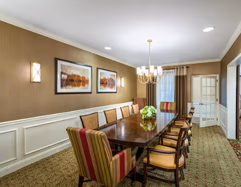 Long wooden dining table with upholstered chairs beneath a chandelier in an elegantly decorated dining room with framed artwork and patterned carpet.