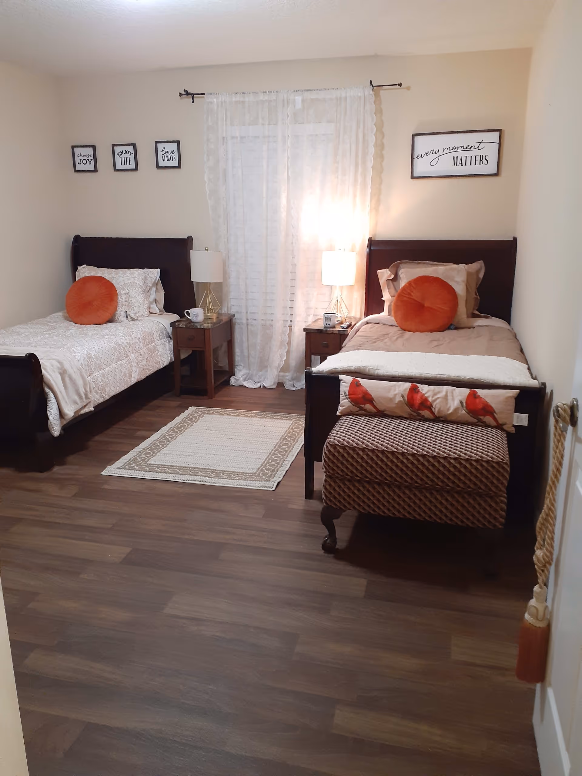 A cozy bedroom with two single beds, each with a dark wooden frame, beige bedding, and an orange round pillow. Between the beds are two wooden nightstands with lamps and mugs. Sheer white curtains cover the window in the center of the back wall. The walls are decorated with framed inspirational quotes. A small patterned bench with a pillow featuring red birds is at the foot of the right bed. The floor is wood with a small beige and white rug in the center.