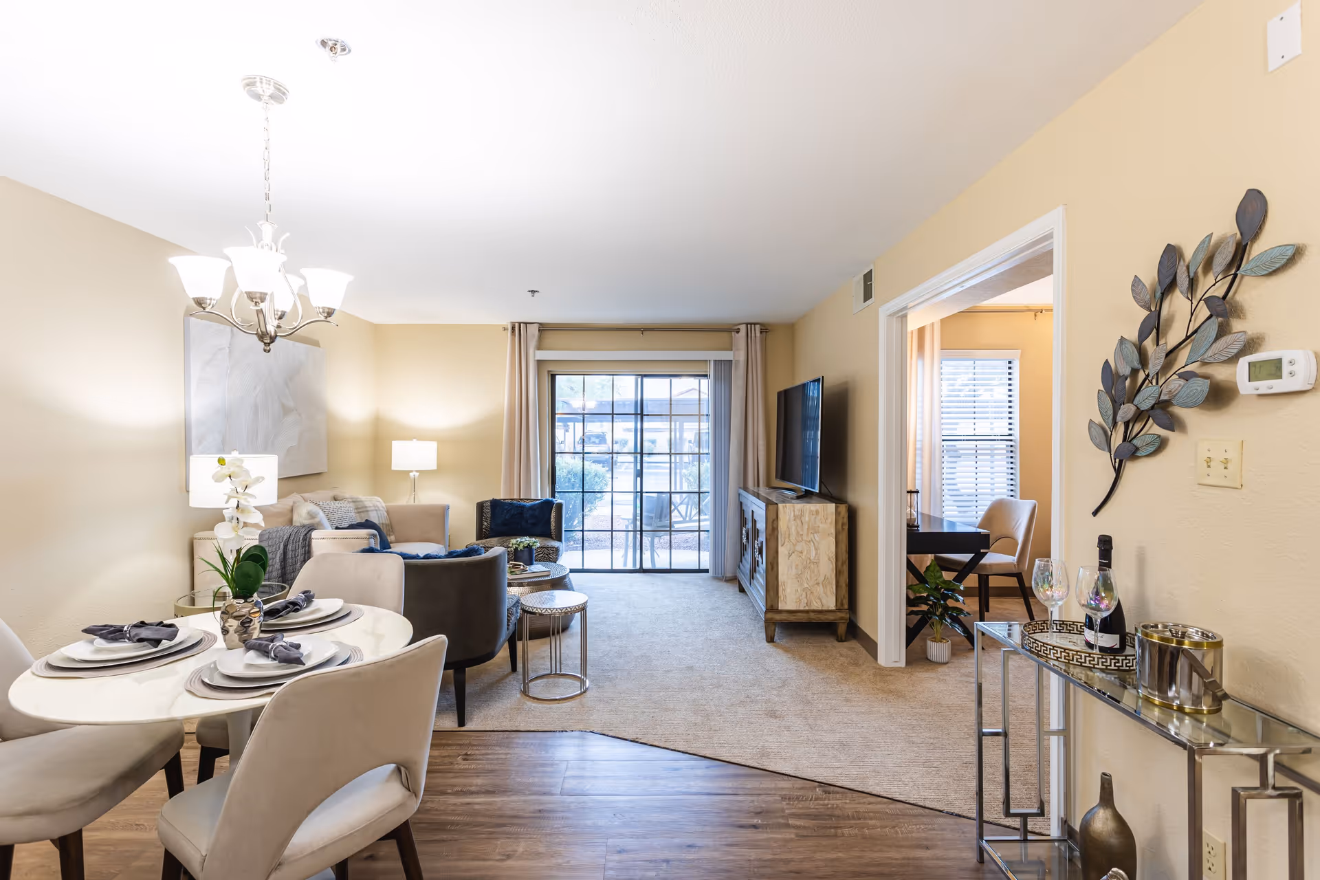 A bright and cozy living and dining area in a senior living facility. The dining area features a round table set with plates, napkins, and a small plant centerpiece, surrounded by four upholstered chairs. The living area has a sofa, armchair, side tables, a floor lamp, and a TV mounted on a wooden cabinet. Large sliding glass doors with curtains lead to an outdoor space. A small glass console table with decorative items and a bottle of wine is positioned against the wall near an open doorway leading to another room with a desk and chair. The walls are painted beige, and the floor transitions from wood in the dining area to carpet in the living area.