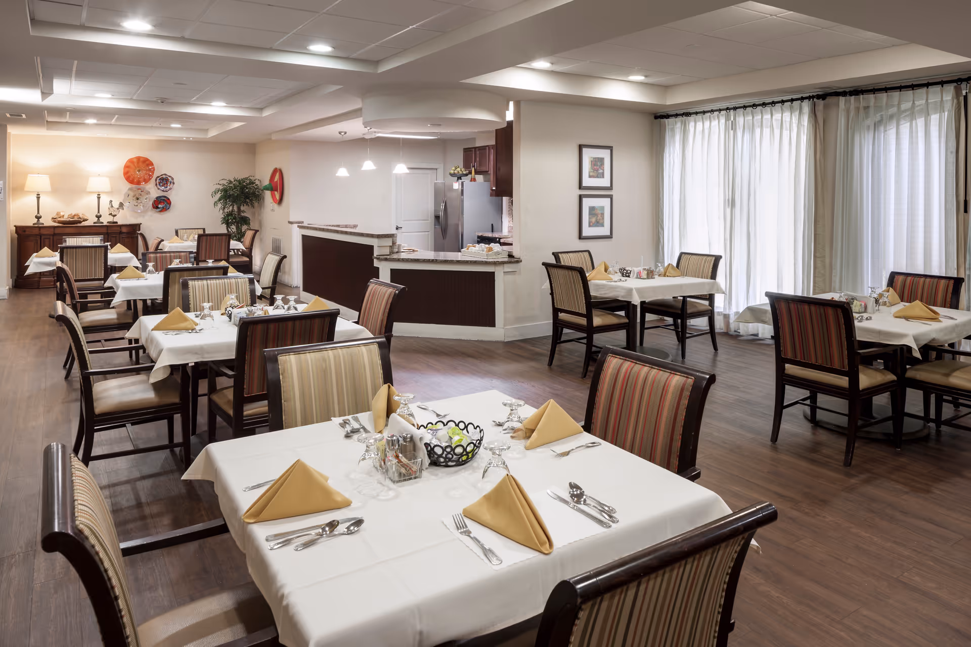 A dining room with several tables covered in white tablecloths, each set with folded beige napkins, silverware, and glassware. The room has wooden flooring, striped upholstered chairs, large windows with sheer curtains, and a counter area with pendant lights in the background. Decorative wall art and lamps are also visible.