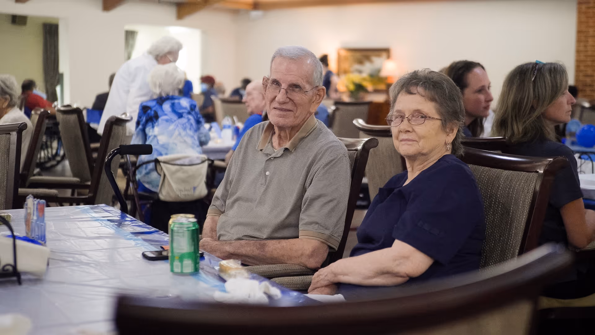 An elderly man and woman sitting together at a table in a dining area with other people in the background. The man is wearing glasses and a striped polo shirt, and the woman is wearing glasses and a dark blue top. There are drinks and napkins on the table, and the room has a warm, communal atmosphere.
