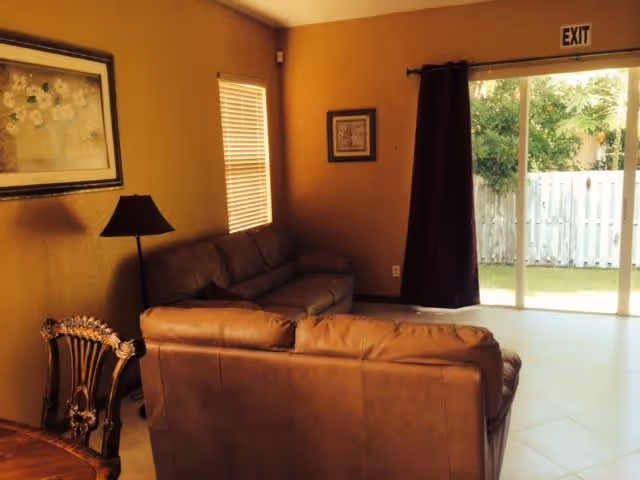 A cozy living room with two brown leather sofas, a wooden chair, a floor lamp, and framed artwork on the walls. There is a window with blinds and a sliding glass door with a dark curtain leading to an outdoor area with a white fence and greenery. An exit sign is visible above the sliding door.