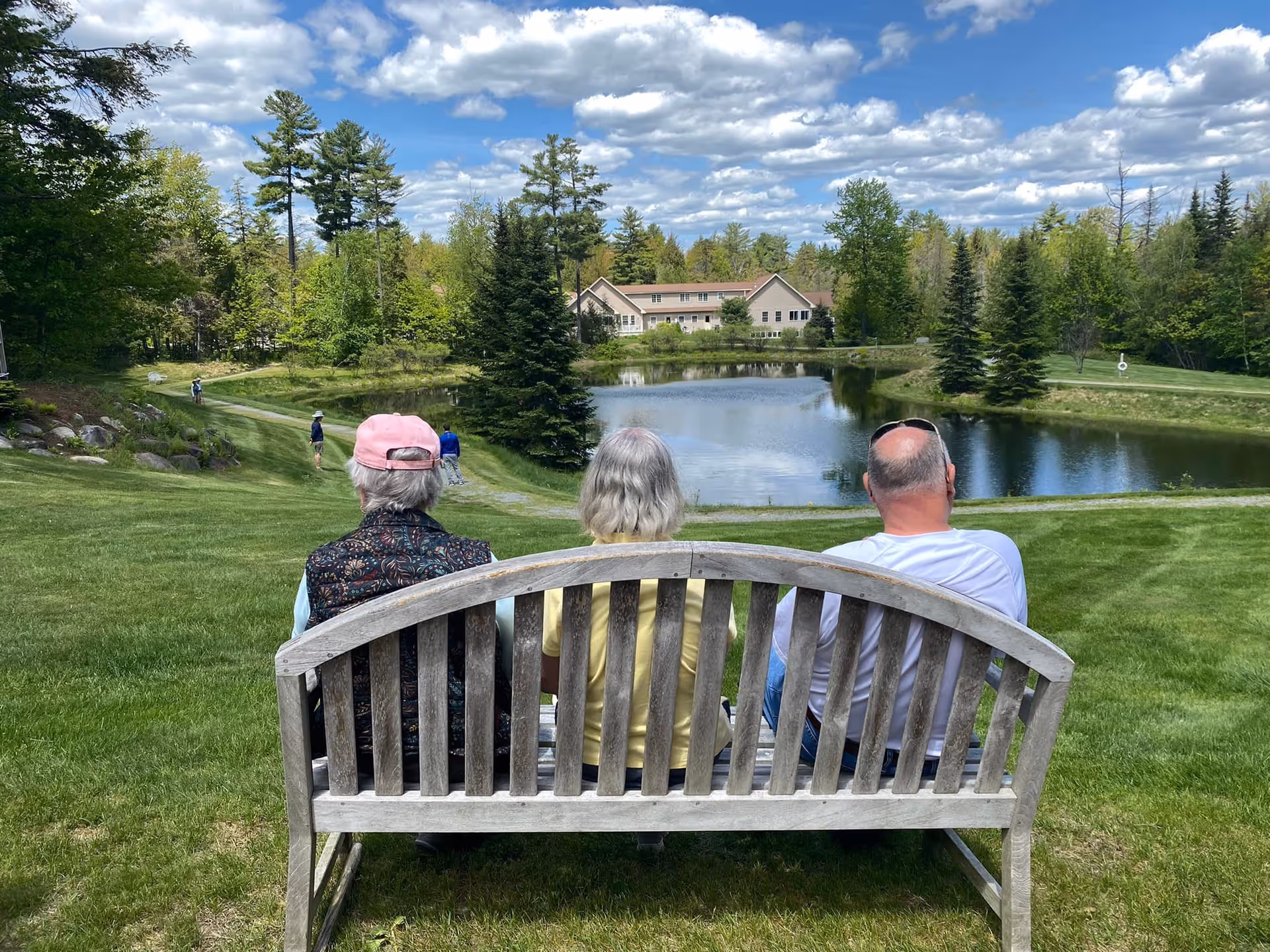 Three people sitting on a wooden bench facing a pond surrounded by green grass and trees under a partly cloudy sky. In the background, there is a large building and a few people walking along a path near the pond.