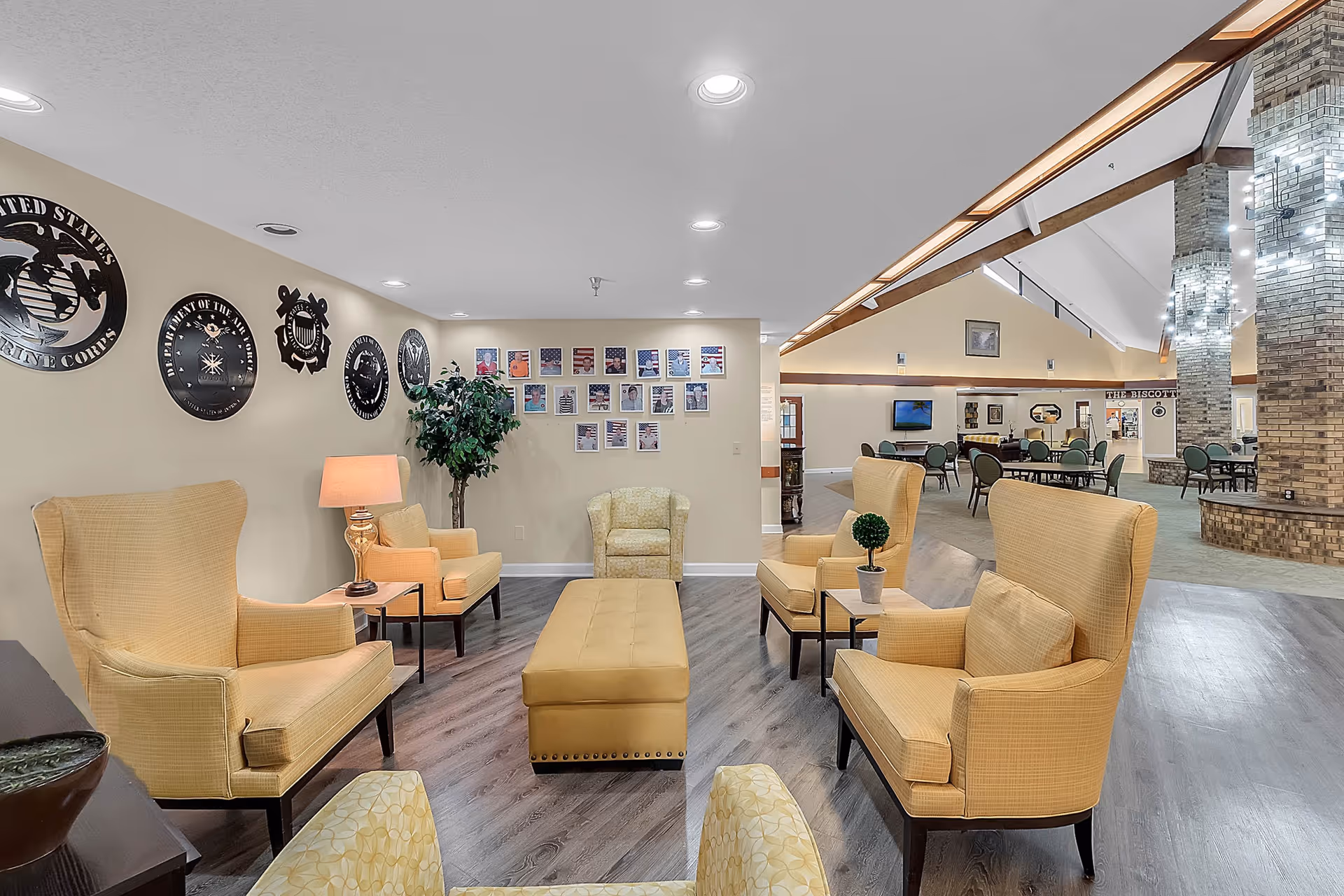 A spacious senior living facility common area with yellow upholstered armchairs and a matching ottoman arranged around small tables. The walls display military emblems and framed photos of veterans. The room features wood flooring, a high ceiling with wooden beams, and a brick column decorated with string lights. In the background, there are additional tables and chairs, a television, and a sign that reads 'THE BISCOTTI'.