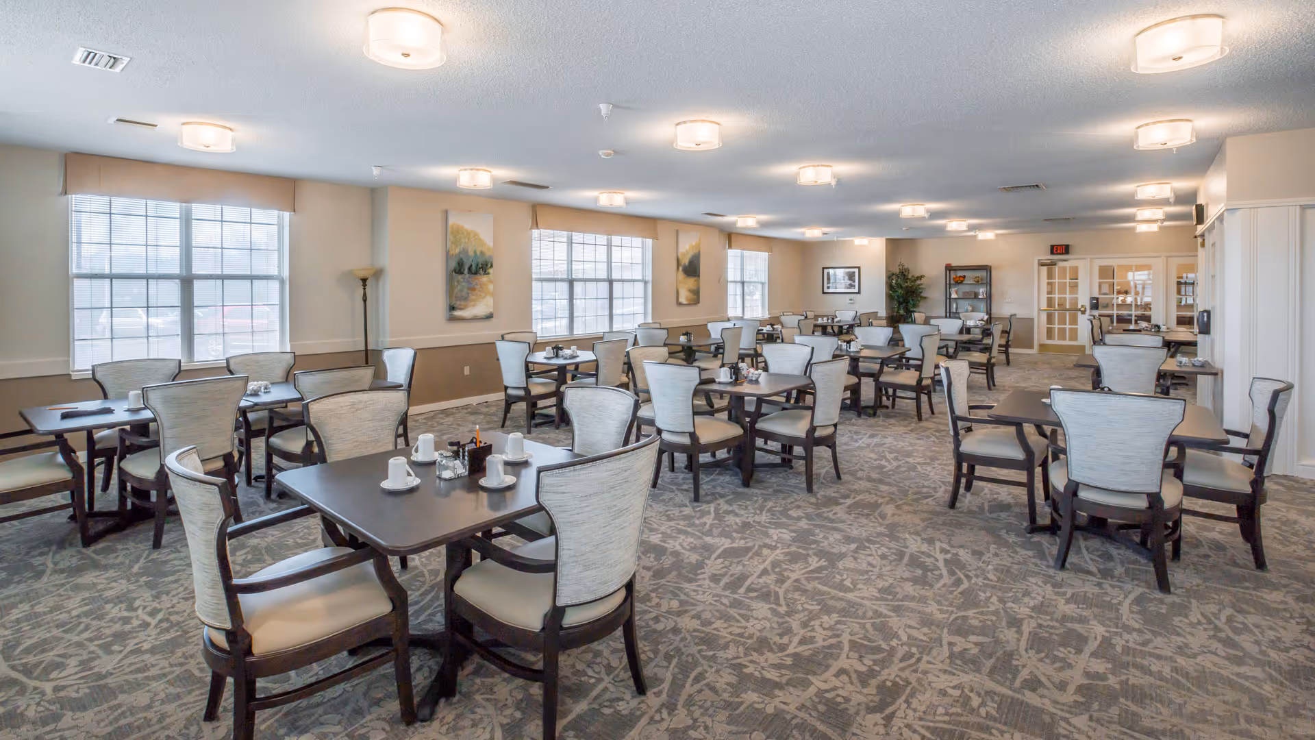 Large, bright communal dining room with multiple set tables and upholstered chairs on patterned carpet.