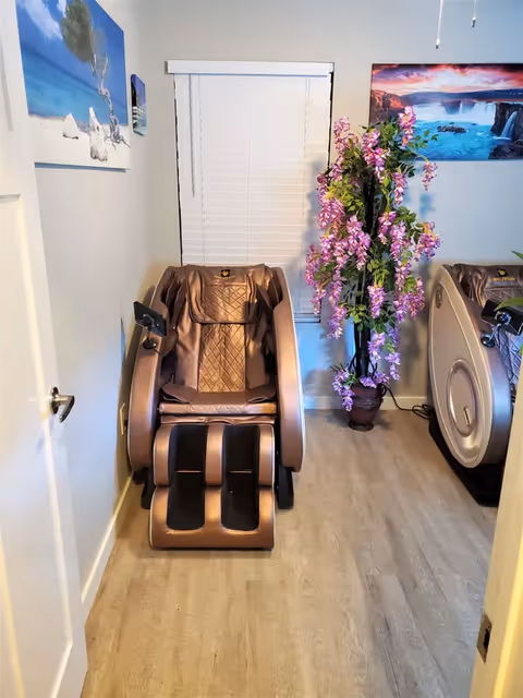 A small room with a brown massage chair positioned in front of a window with closed white blinds. To the right of the chair is a tall potted plant with purple flowers. The walls are decorated with two landscape paintings, one showing a beach scene and the other a waterfall. The floor is light-colored wood, and there is a door partially visible on the left side.