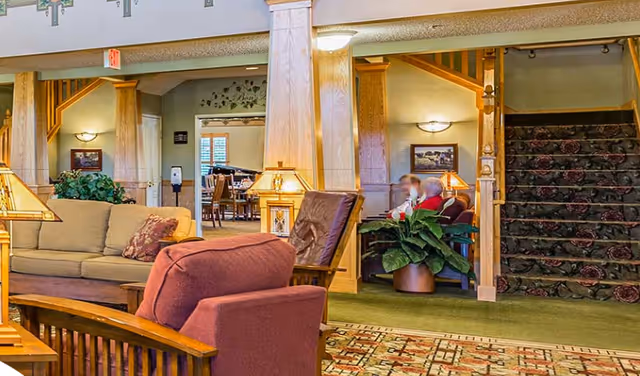 A cozy senior living facility common area with comfortable seating including a beige sofa and a red armchair. There are wooden pillars and a staircase with floral carpet leading upstairs. Two people are seated and conversing near a large potted plant. The room is warmly lit with table lamps and wall sconces, and a dining area is visible in the background.