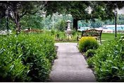 Paved garden path leading to a bench and decorative fountain with a lake and trees in the background.