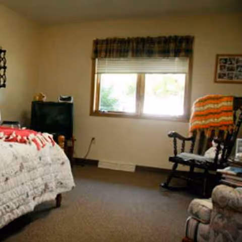 A bedroom in a senior living facility with a quilted bed, window, rocking chair, and dresser.
