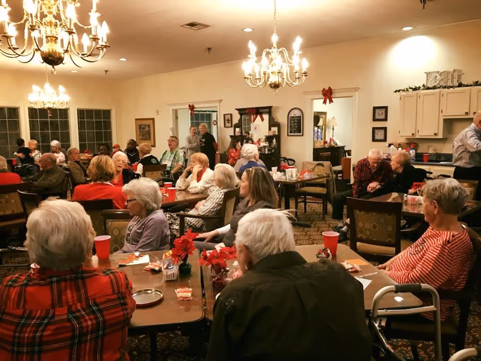 A large group of elderly people gathered in a well-lit dining room with chandeliers, sitting at tables decorated with red poinsettias and holiday-themed items. Some people are engaged in conversation, and the room has a warm, festive atmosphere with holiday decorations such as red bows on door frames. The background shows a kitchen area and a cabinet with various items.
