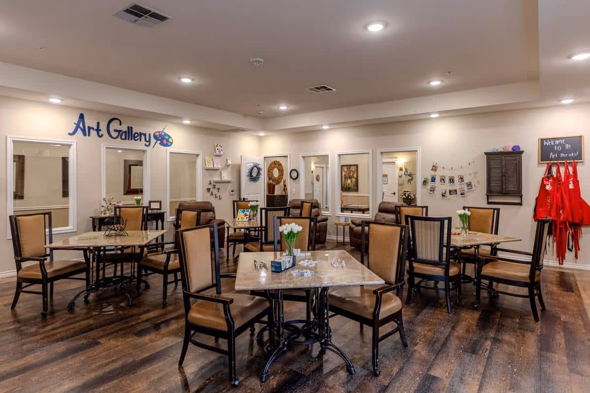 A well-lit room with several tables and chairs arranged neatly on a wooden floor. The walls are decorated with art pieces and a sign that reads 'Art Gallery' with a painter's palette graphic. There are red aprons hanging on the right side under a small chalkboard sign that says 'Welcome to Art throb!'. The room appears to be a communal space for art activities.
