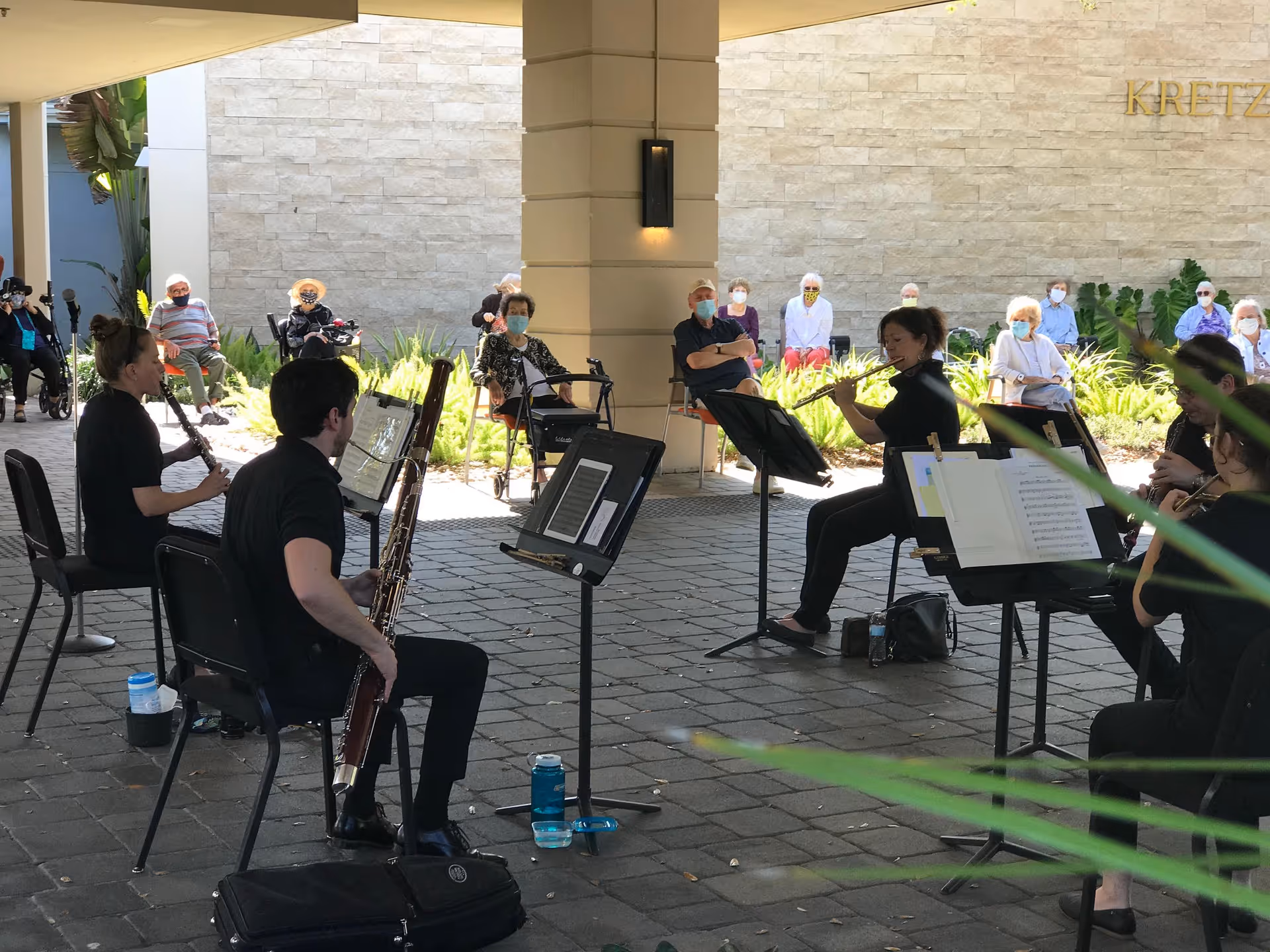 A small group of musicians playing woodwind instruments seated in a semi-circle under a covered outdoor area, performing for an audience of elderly people who are seated socially distanced and wearing masks in front of a stone wall with the word 'KRETZ' partially visible.
