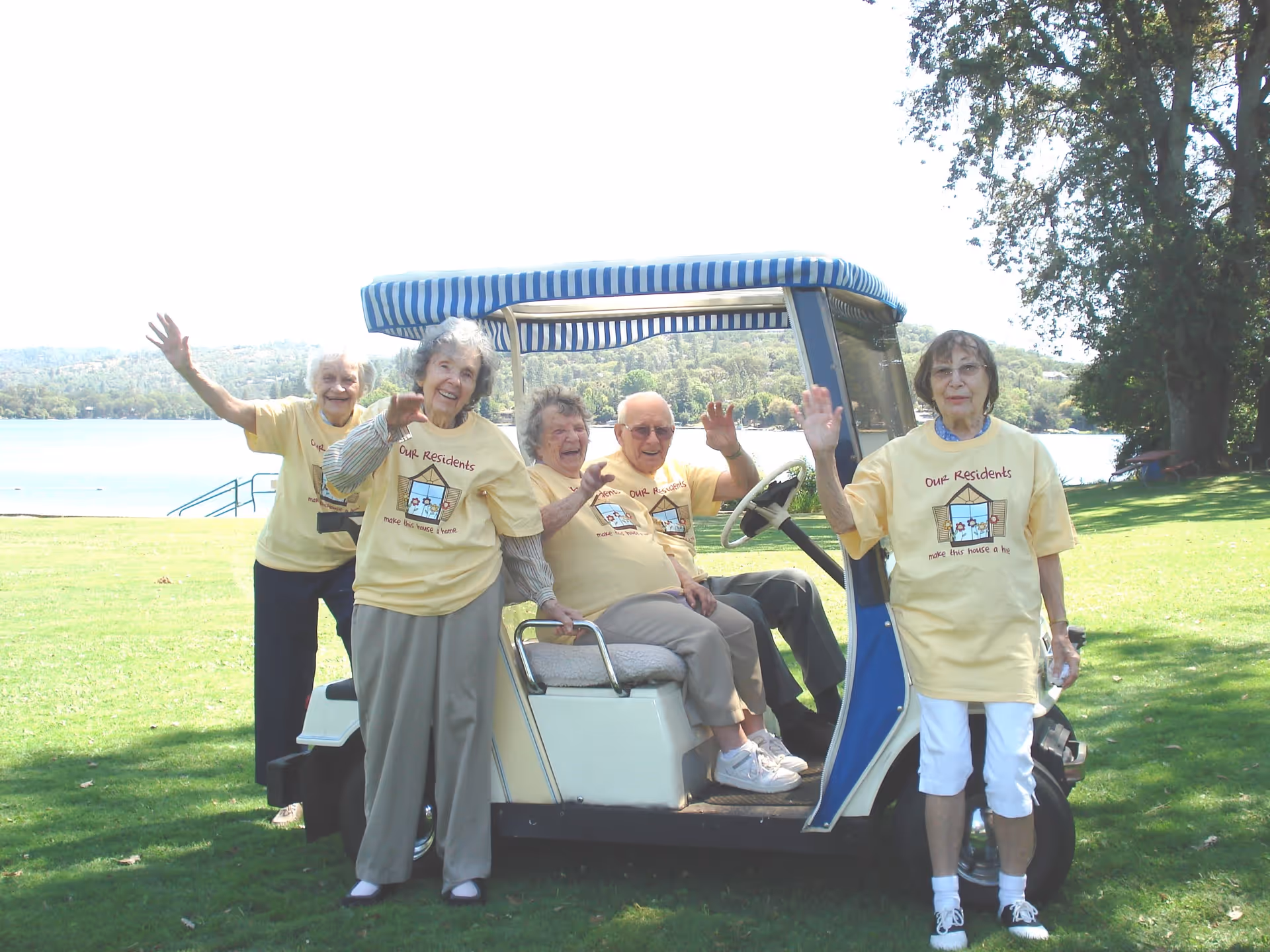 Five elderly individuals wearing matching yellow t-shirts are smiling and waving while gathered around and sitting in a golf cart on a grassy area near a body of water with trees in the background.