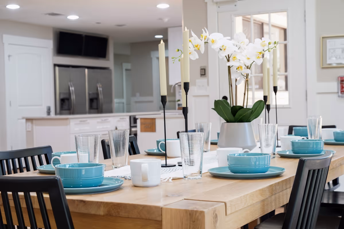 A dining table set with turquoise plates, bowls, white mugs, and clear drinking glasses. The table also features a white orchid centerpiece and tall white candles in black holders. In the background, there is a modern kitchen with stainless steel refrigerators and white cabinetry.