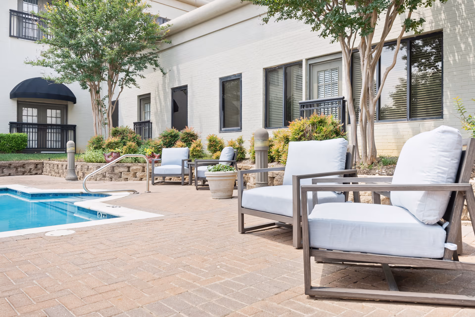 Outdoor patio area with cushioned lounge chairs arranged near a swimming pool. The area is paved with bricks and surrounded by greenery and trees. The building exterior with windows and doors is visible in the background.