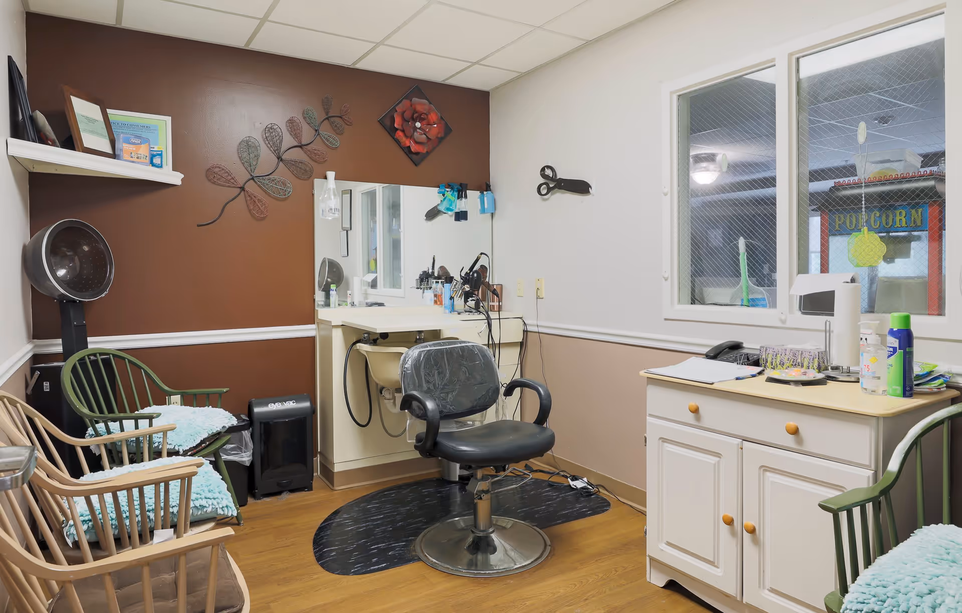Interior of a small salon or beauty care room with a black salon chair in front of a mirror and counter with hair care tools. There are two wooden chairs with green cushions and a hair dryer hood on the left side. A white cabinet with various items on top is on the right side, and a window shows a popcorn machine outside the room. The walls are painted brown and beige with decorative wall art.