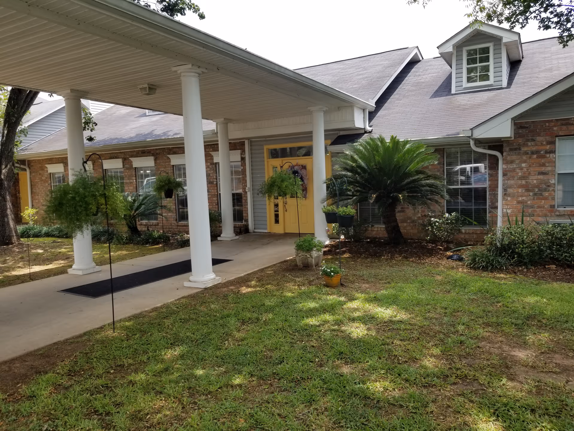 Front exterior view of a single-story brick building with a covered entrance supported by white columns. The entrance has double yellow doors decorated with a wreath. There are several windows along the building, and the surrounding area has green grass, shrubs, and hanging plants.