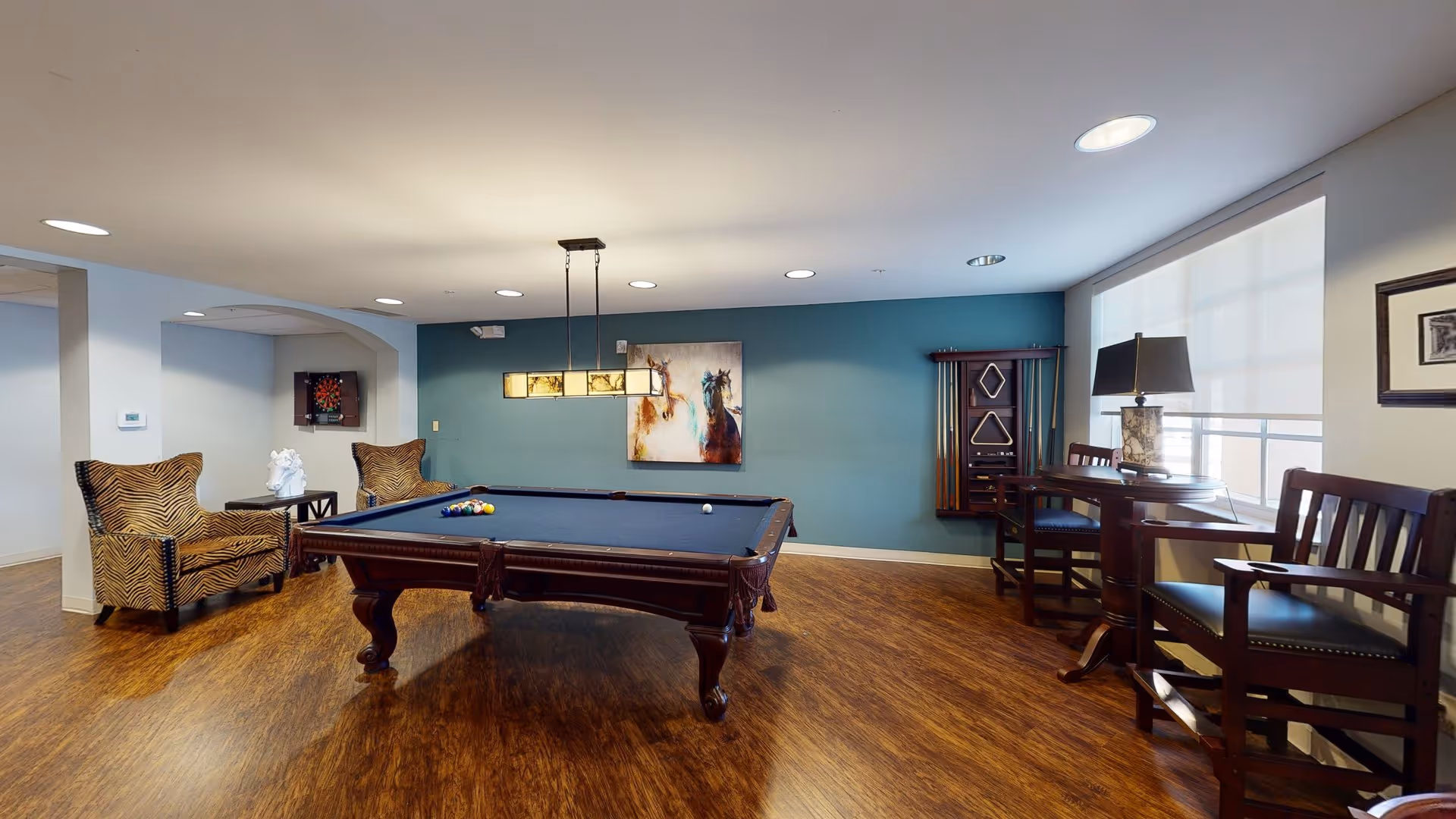 A recreational room with a pool table in the center, two zebra-patterned armchairs on the left, a dartboard mounted on the wall, a painting of two horses on a teal accent wall, a wall-mounted pool cue rack, and a small table with two chairs and a lamp near a large window with blinds.