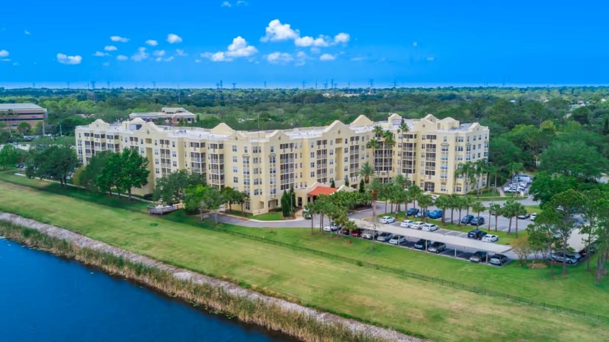 Aerial view of Stratford Court of Palm Harbor, a large multi-story residential building surrounded by green lawns, trees, and a parking lot, with a body of water in the foreground and a clear blue sky above.
