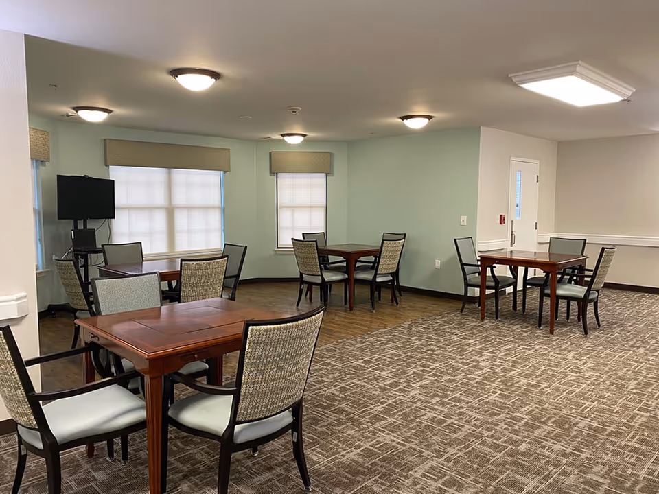 A senior living facility common area with multiple wooden tables and cushioned chairs arranged on a carpeted floor. The walls are painted light green and white, with windows covered by beige blinds. Ceiling lights illuminate the room, and a television is mounted on a stand in the corner.