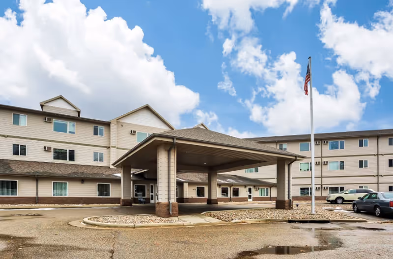 Exterior view of Minot Lodge, a three-story senior living facility with beige siding and brown brick accents. The building features a covered entrance with a peaked roof, a flagpole with an American flag, and a parking area with several cars. The sky is partly cloudy with blue patches.