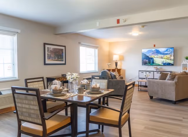 A bright and spacious living and dining area in a senior living facility. The dining area features a wooden table set for four with plates, bowls, cups, and napkins. The living area has two sofas, a coffee table, a side table with a lamp, and a flat-screen TV mounted on the wall displaying a scenic mountain landscape. The room has large windows with blinds, light-colored walls, and wood flooring.