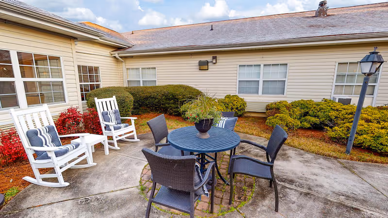 Outdoor patio area at PruittPlace - Wesleyan with a round table surrounded by four black wicker chairs and a potted plant centerpiece. Two white rocking chairs with blue and white striped cushions are placed nearby. The patio is surrounded by bushes and the exterior walls of the building with several windows.
