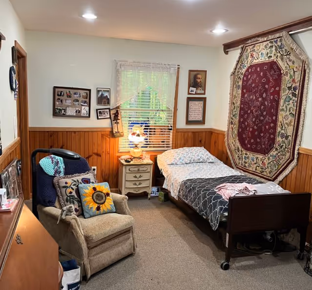 A cozy bedroom in an assisted living facility featuring a single bed with patterned bedding, a wooden nightstand with a lamp, a window with blinds and lace curtains, and a comfortable armchair with colorful pillows including a sunflower design. The walls are half-paneled with wood and decorated with framed pictures and a large decorative tapestry.