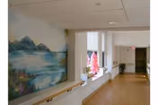 Bright interior hallway in a senior living facility with a landscape mural on the wall, handrail, windows with decorations, and wood flooring.