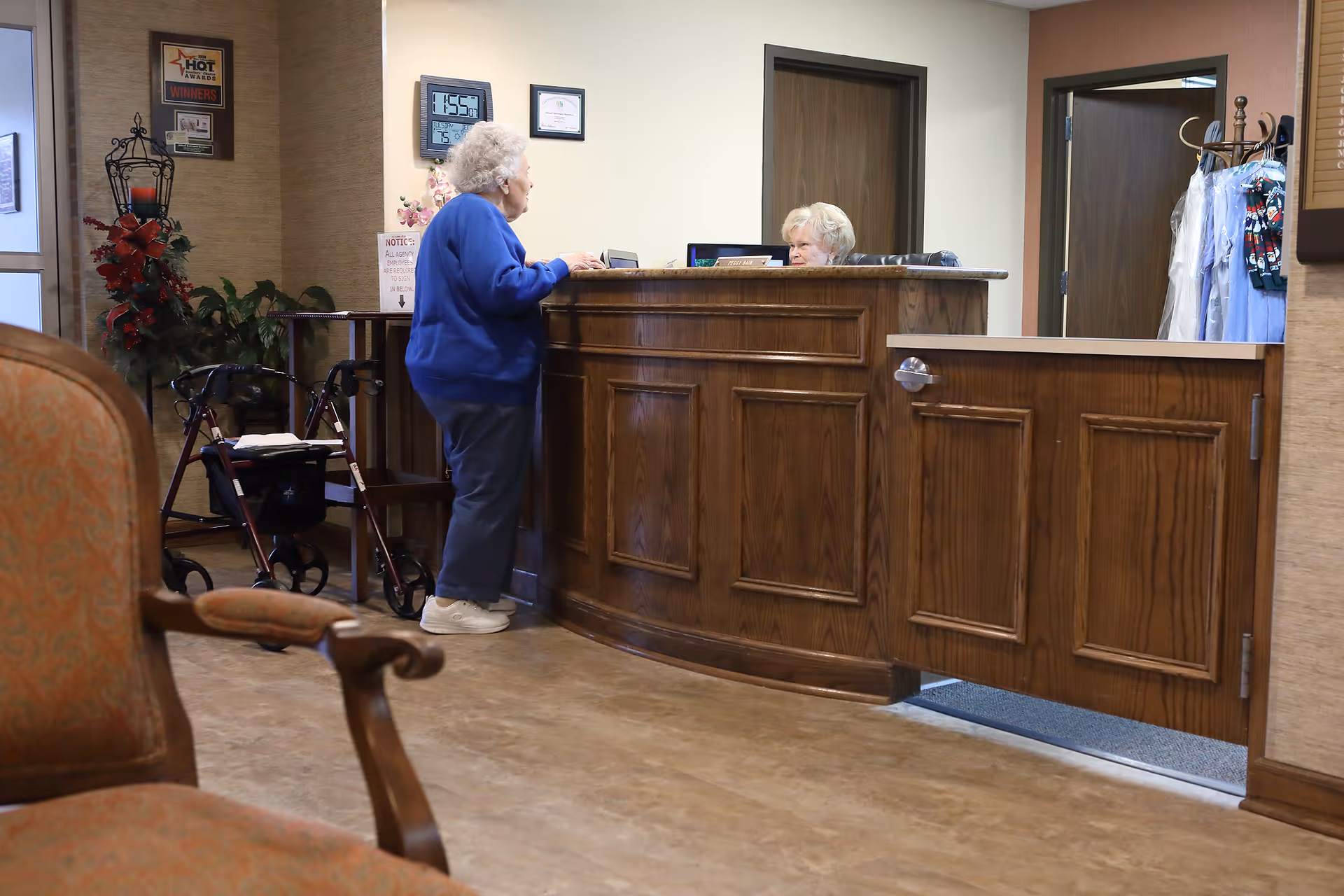 An elderly woman in a blue sweater stands at a wooden reception desk talking to a receptionist seated behind the desk. A walker is parked nearby, and there are chairs and plants in the background. The setting appears to be a welcoming indoor area of a senior living facility.